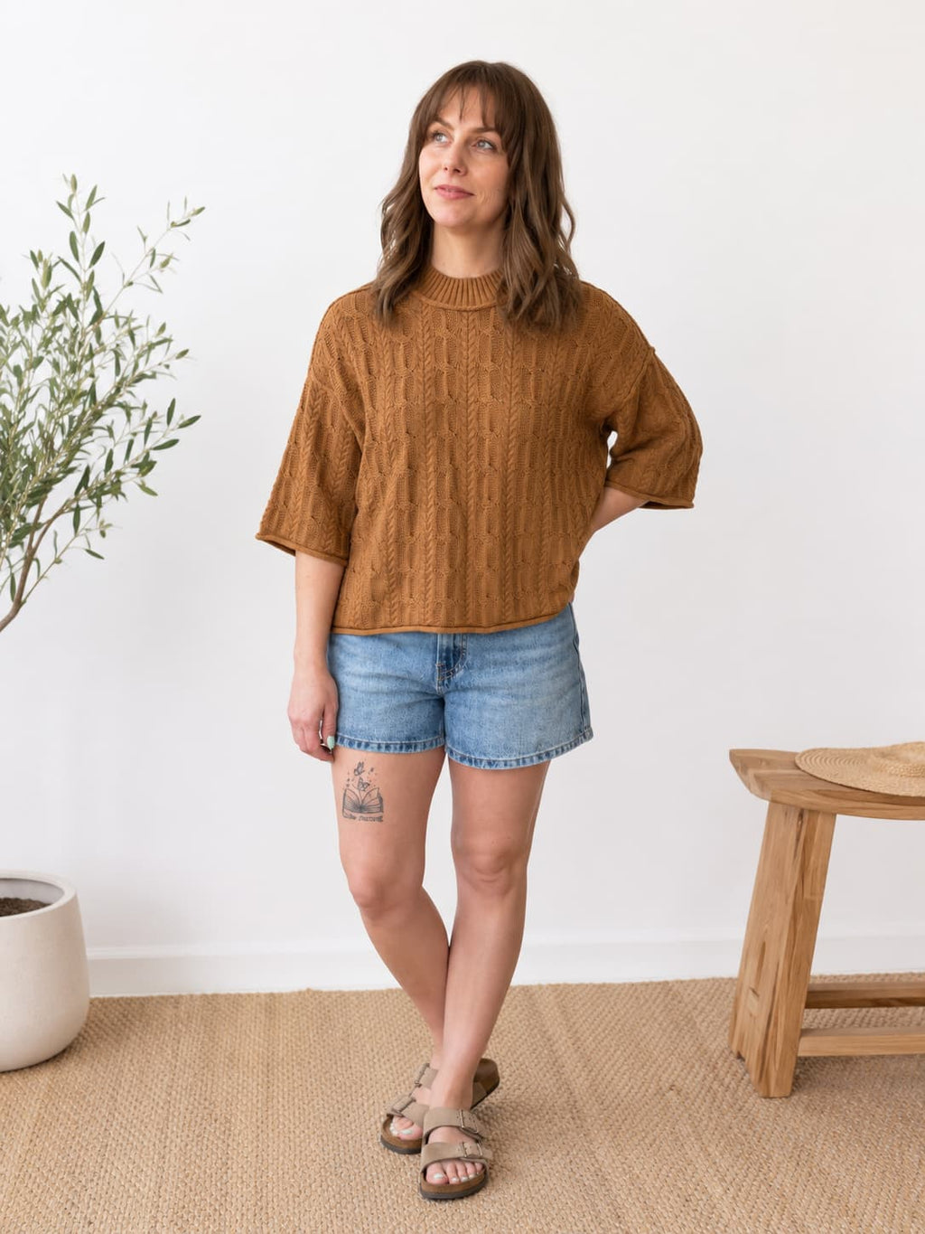 A woman modeling a textured, ribbed brown knit sweater with elbow-length sleeves, paired with denim shorts and sandals, standing in a bright room with a white wall, a potted plant, and a wooden stool.