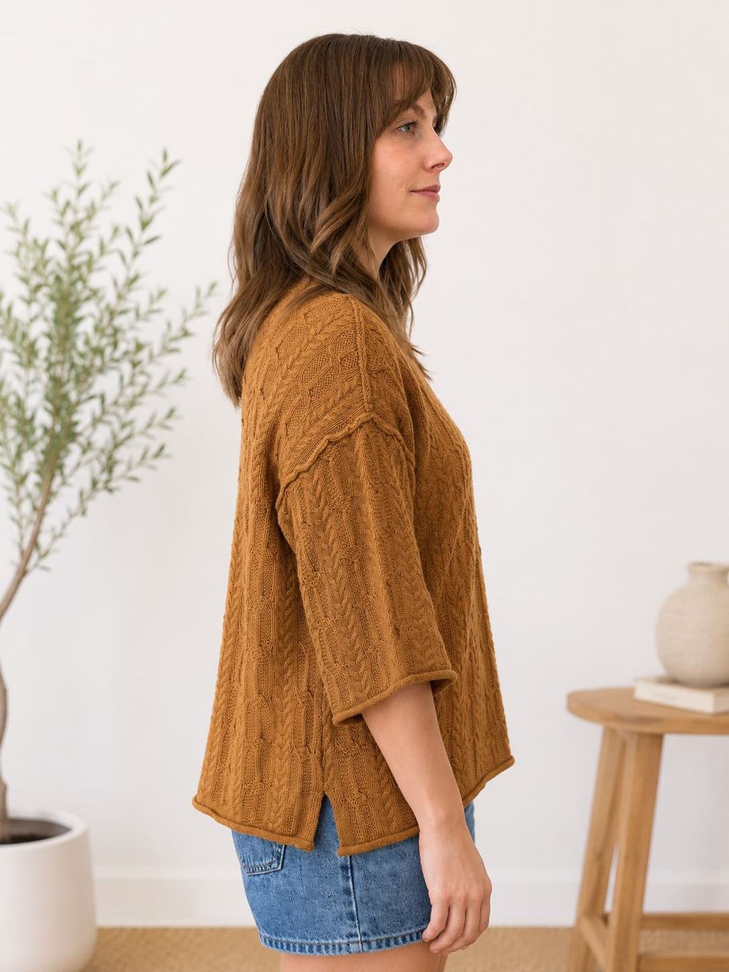A woman wearing a textured, cable-knit mustard-brown short-sleeve top and denim shorts, standing in profile in a bright, modern room with a potted plant and a wooden side table.