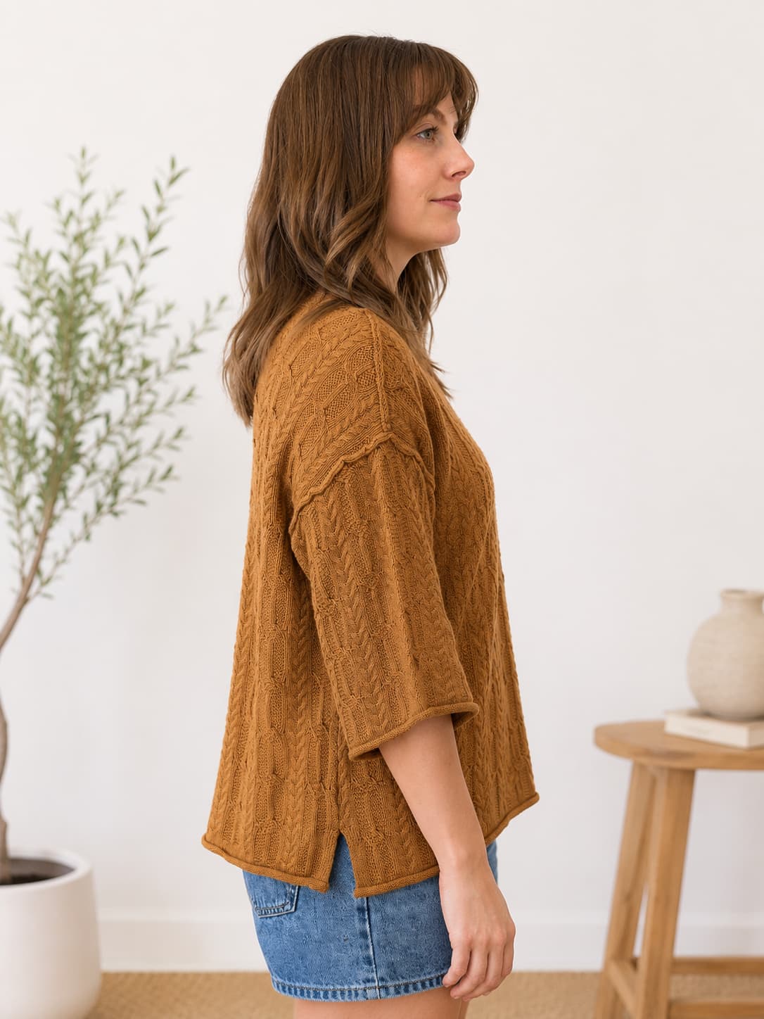 A woman wearing a textured, cable-knit mustard-brown short-sleeve top and denim shorts, standing in profile in a bright, modern room with a potted plant and a wooden side table.