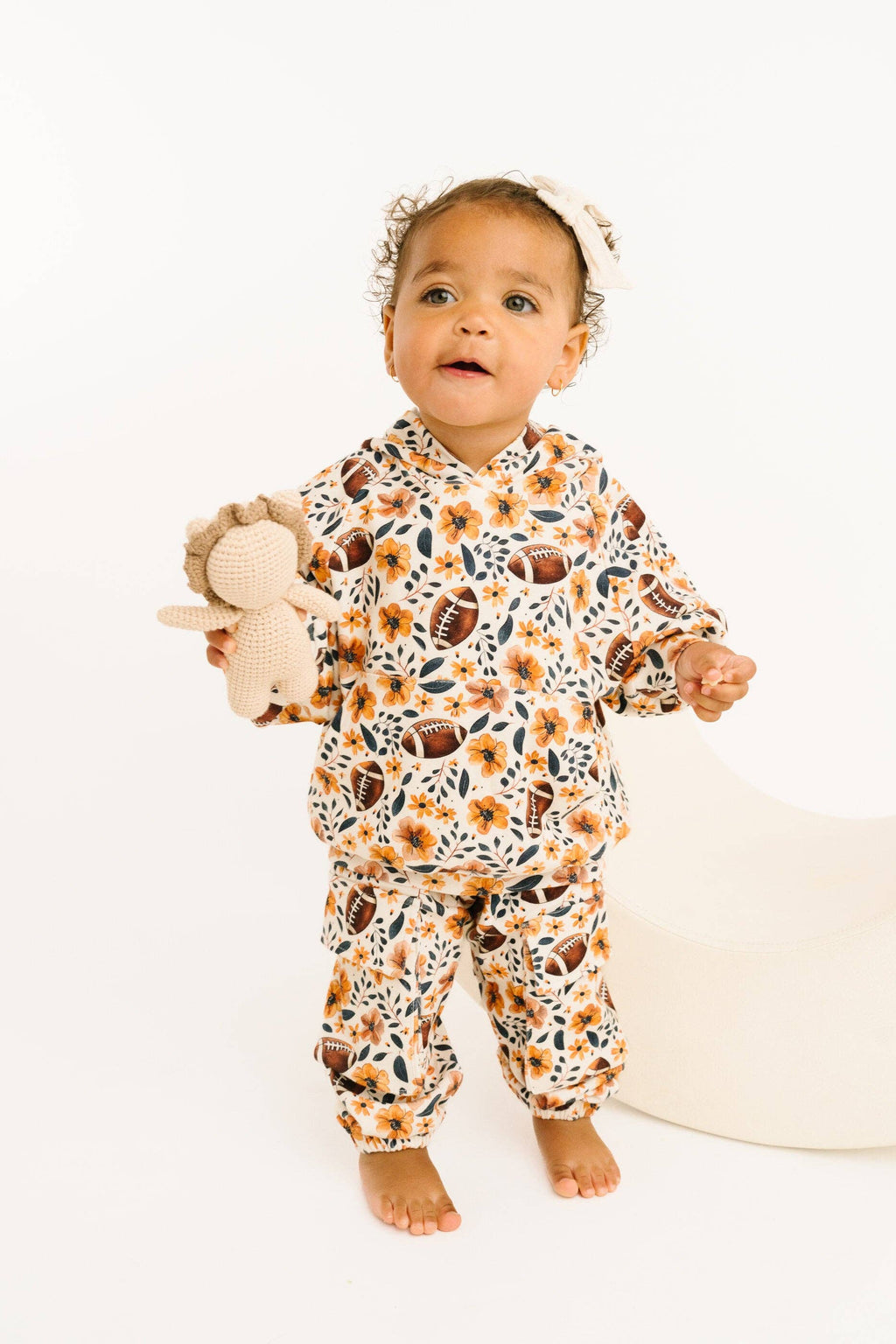 A toddler stands barefoot against a clean white studio background, wearing a matching two-piece outfit with a white base and an allover print of orange flowers, brown footballs, and navy leaves. The child is holding a small beige stuffed animal and wearing a white headband.