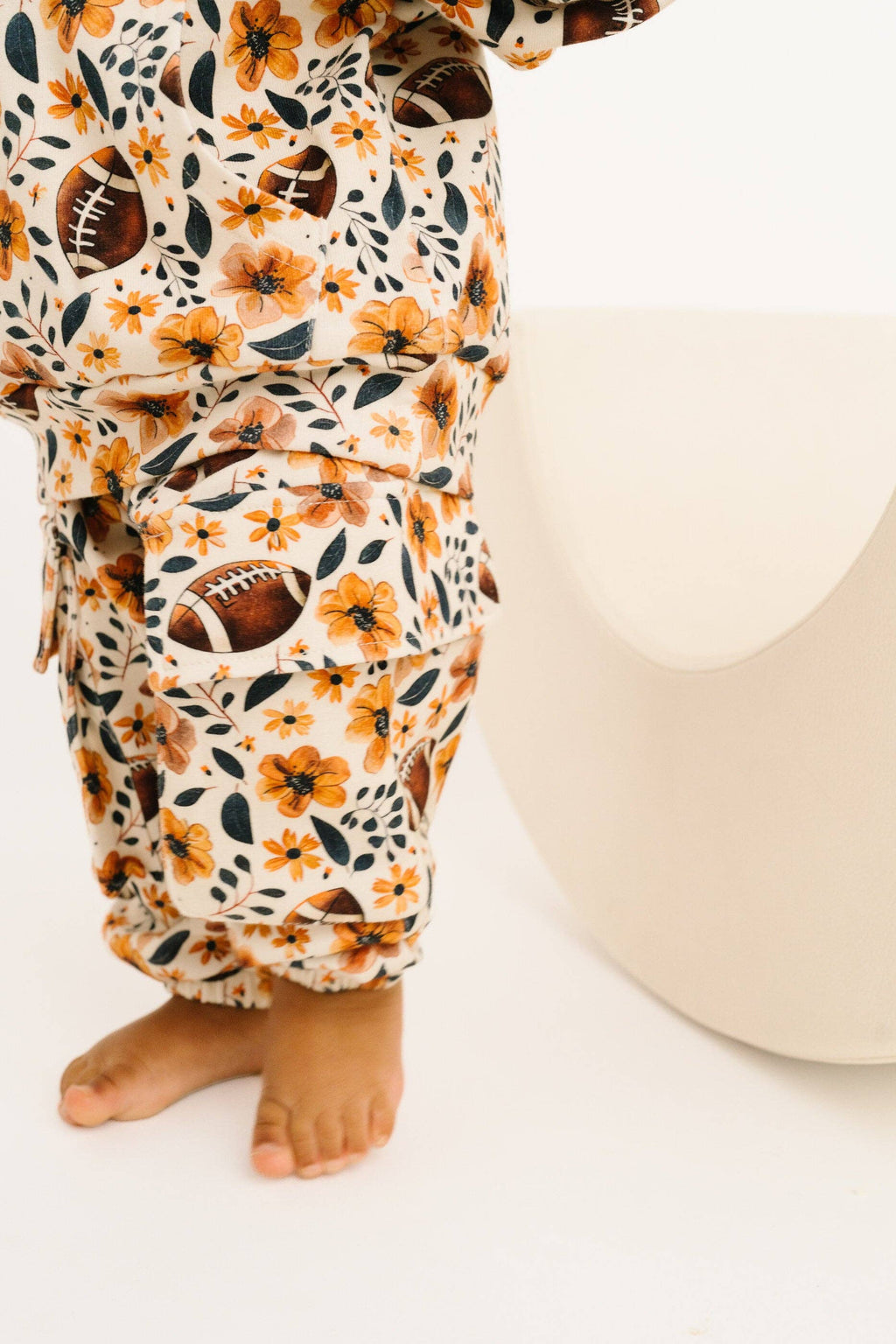 A close-up of a child wearing cream-colored cargo pants with a floral and football print, standing barefoot next to a modern white stool.