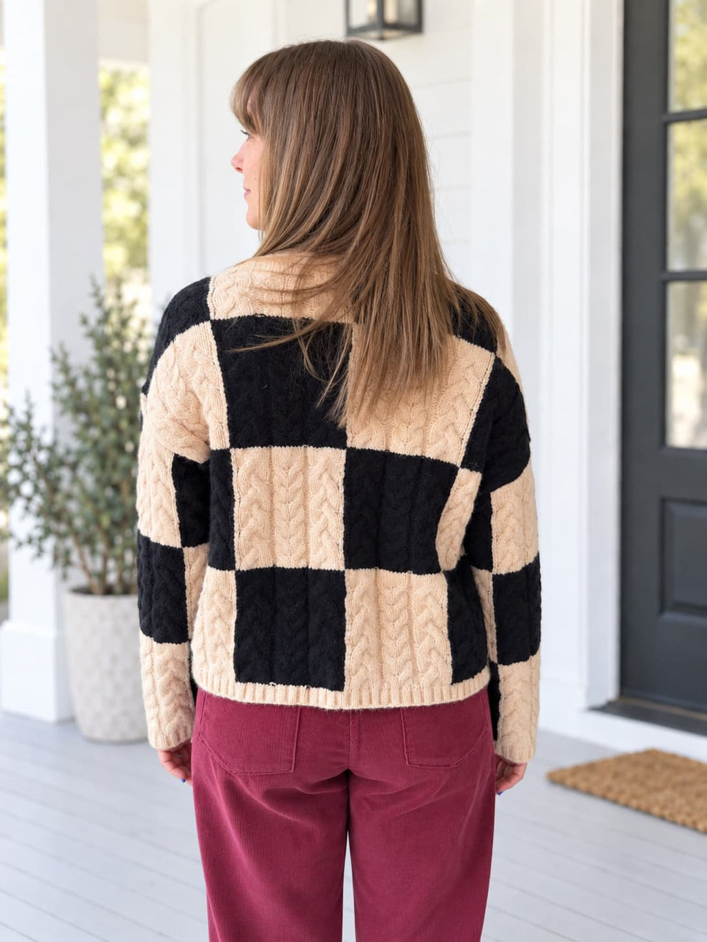 A woman viewed from behind wearing a black and beige checkered cable-knit cardigan and burgundy pants, standing on a white porch.