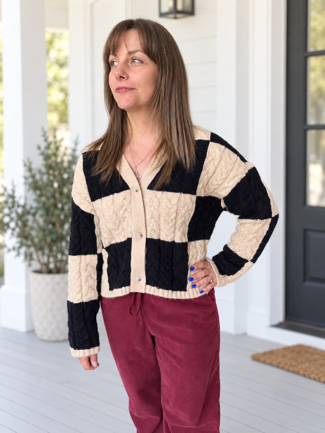 A woman wearing a black and cream cable-knit cardigan with burgundy pants, standing on a white porch.