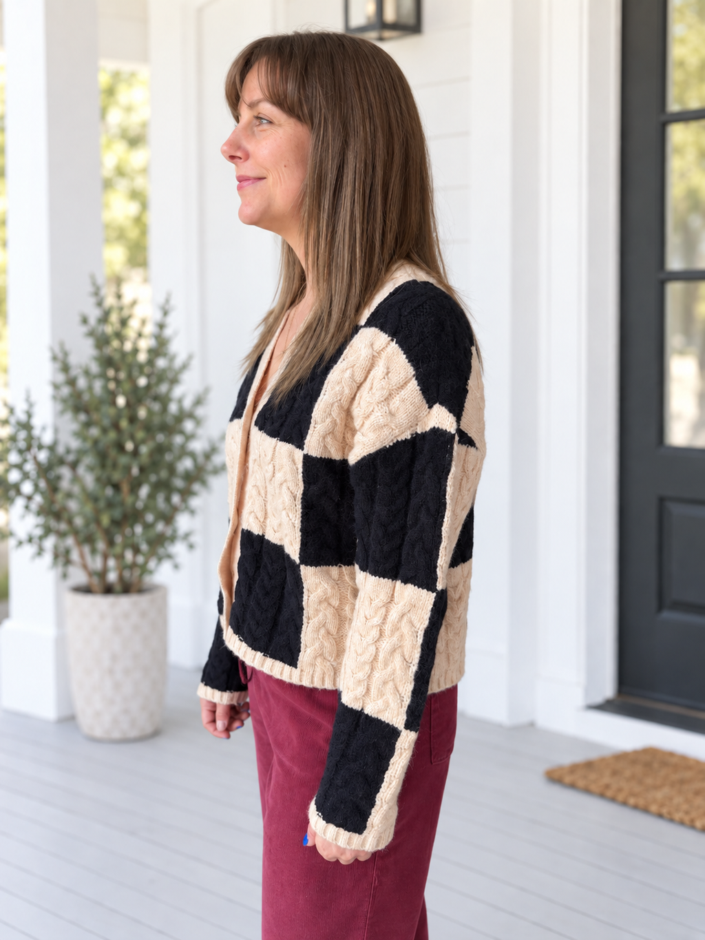 A woman wearing a black and cream checkered cable-knit cardigan sweater and burgundy pants, standing on a white porch.