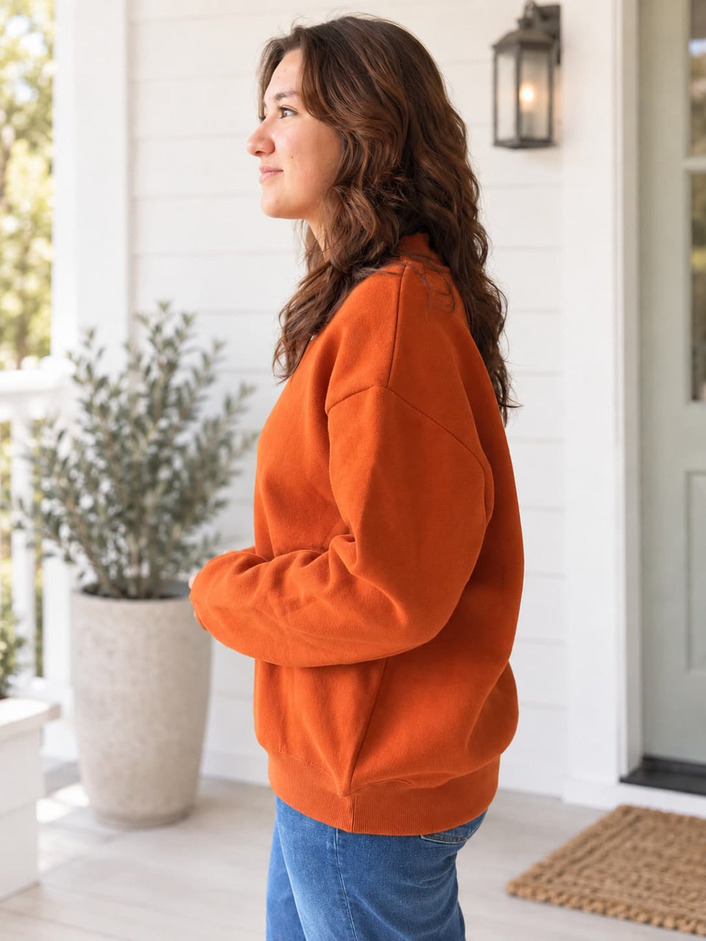 A woman wearing a bright orange crewneck sweatshirt and blue jeans, standing in profile on a white shiplap porch with a potted plant and wall lantern in the background.