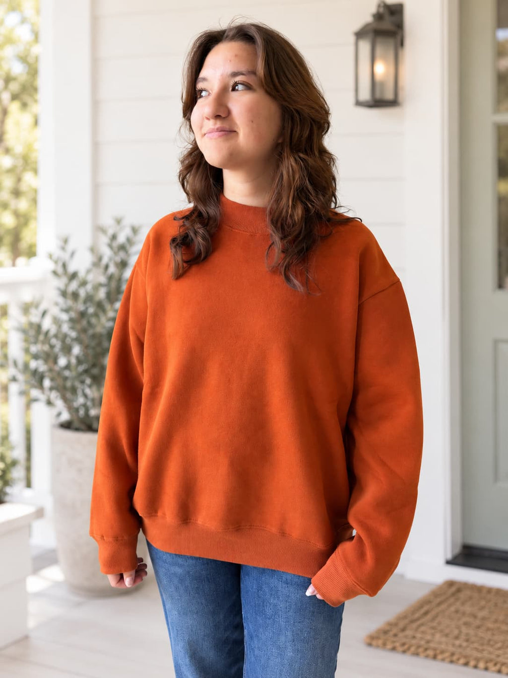 A woman wearing an oversized burnt orange crewneck sweatshirt and blue jeans, standing on a white porch with a potted plant and a lantern in the background.