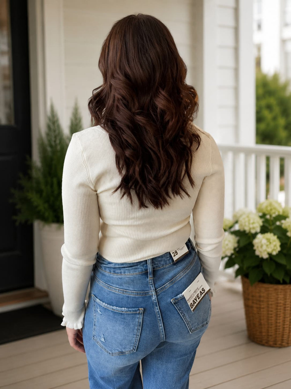 A woman viewed from behind wearing a cream ribbed long-sleeve top and blue jeans, standing on a porch with potted plants in the background.