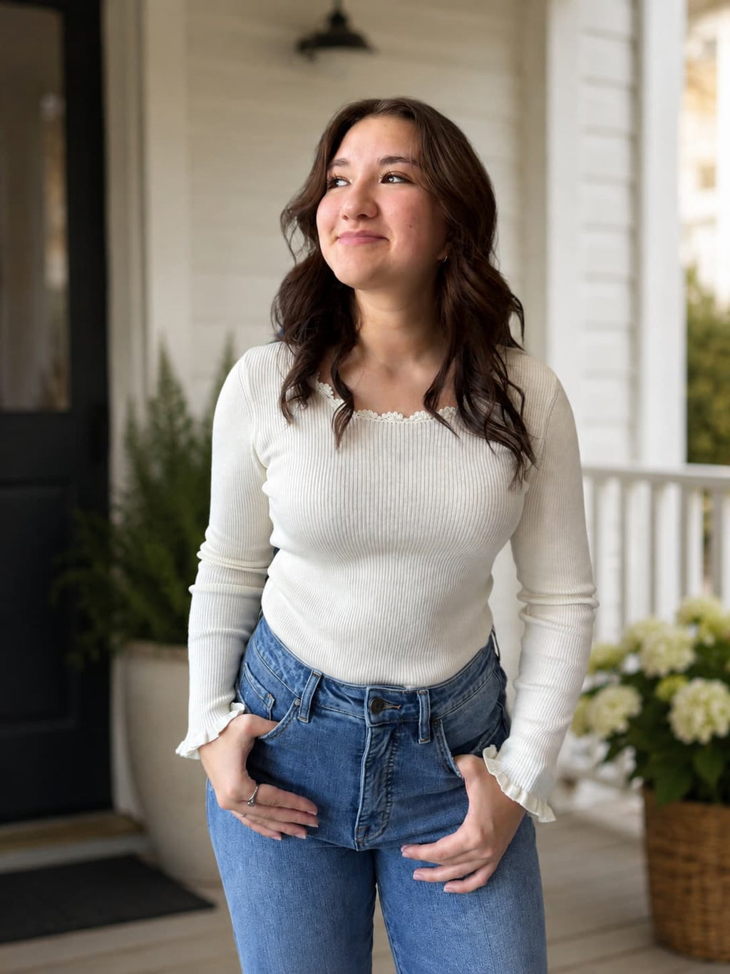 A woman wearing a cream ribbed long-sleeve top with ruffled wrist detail, lace scalloped neckline and blue jeans, standing on a white porch with potted plants in the background.