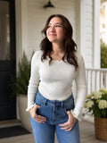 A woman wearing a cream ribbed long-sleeve top with ruffled wrist detail, lace scalloped neckline and blue jeans, standing on a white porch with potted plants in the background.