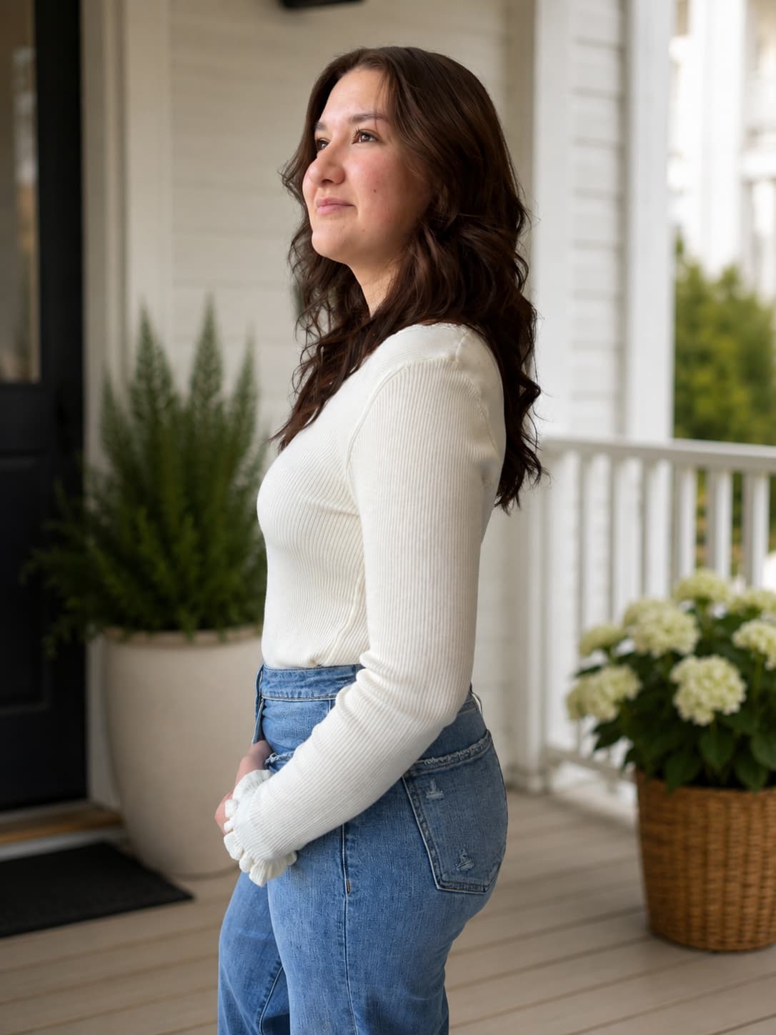 A woman with long brown hair stands in profile on a porch, wearing a fitted white ribbed long-sleeve top with ruffle wrist detail perfect for layering.
