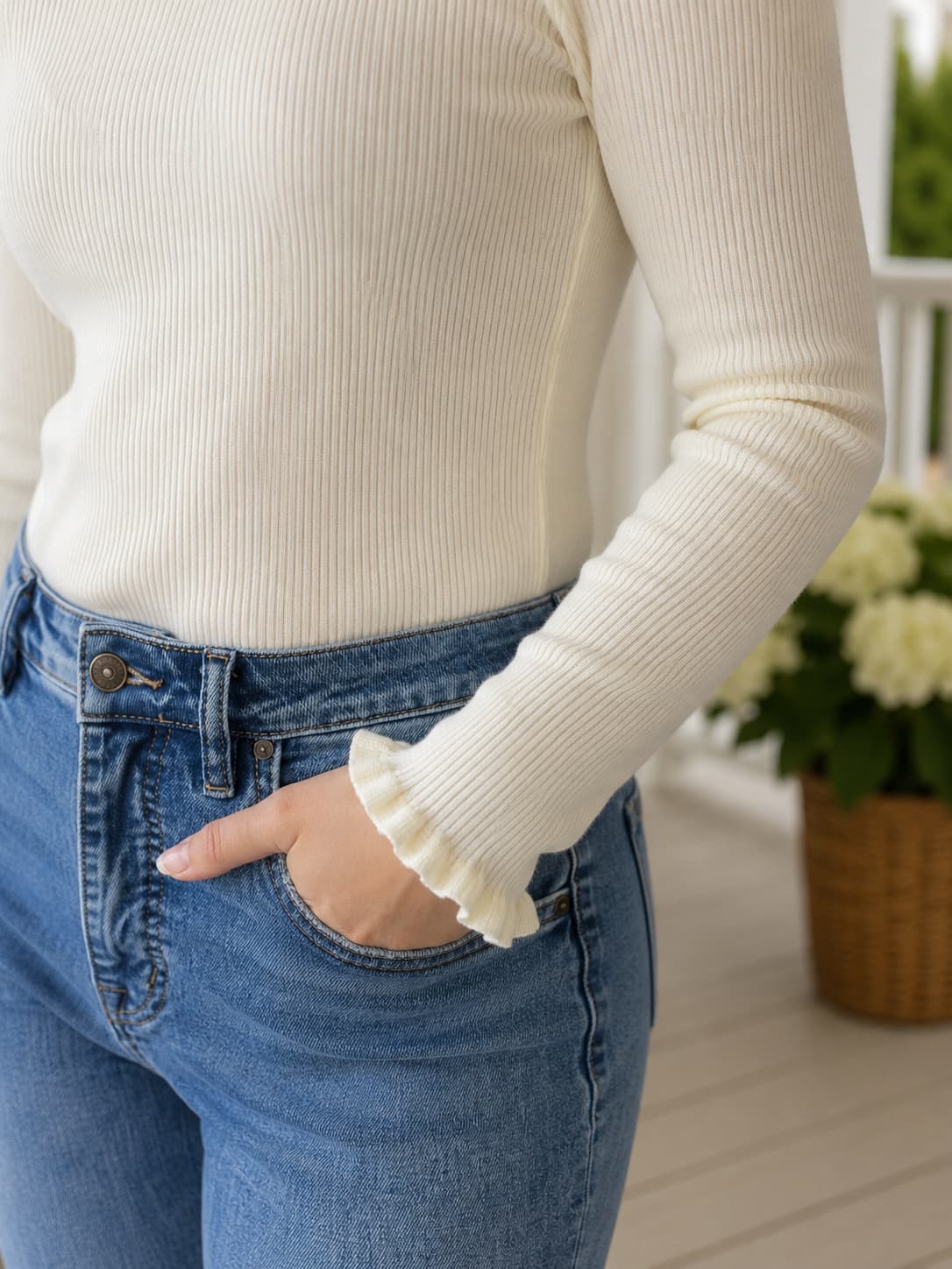A close-up of a person wearing a cream ribbed long-sleeve top with a feminine ruffled wrist detail and blue jeans, standing indoors near a potted plant.