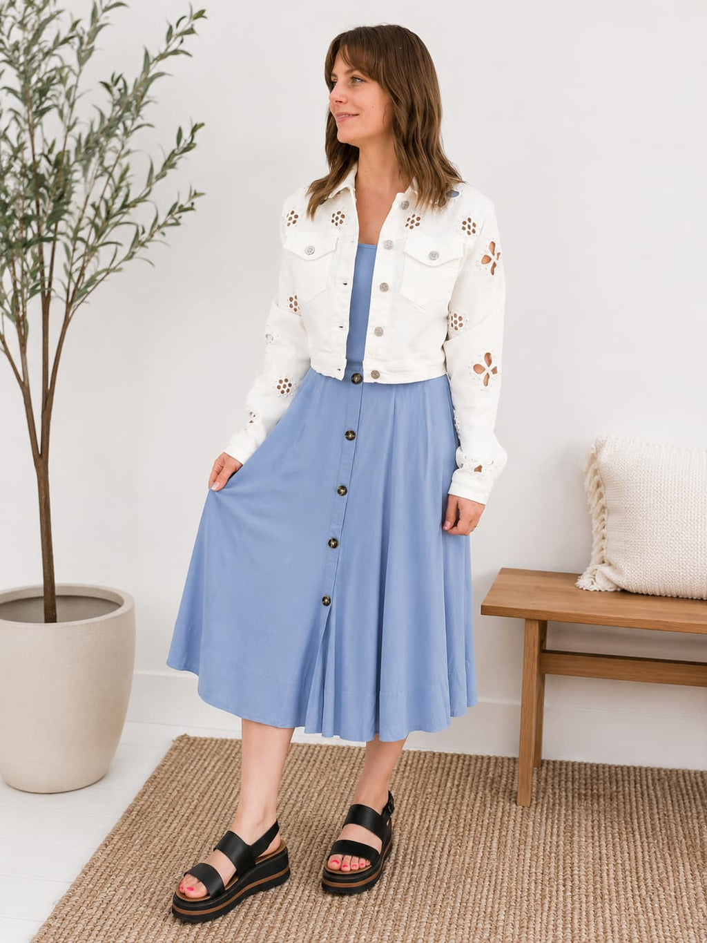 Woman wearing a white denim jacket with embroidered floral patterns and a blue skirt in a room with a plant and bench.