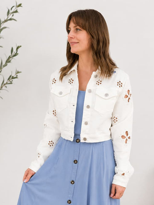 Woman wearing a white denim jacket with floral patterns and a blue skirt in a room with a plant and bench.