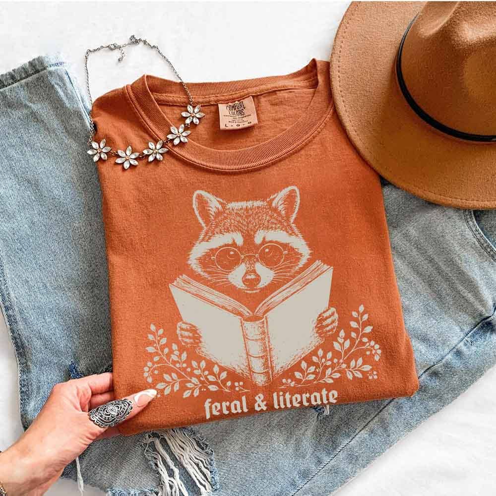 An orange-brown graphic T-shirt laid flat on top of light blue denim jeans, with a tan hat partly visible at the top right and a hand holding the shirt at the bottom left. The shirt features a white raccoon illustration reading a book with the words 'feral & literate' below.