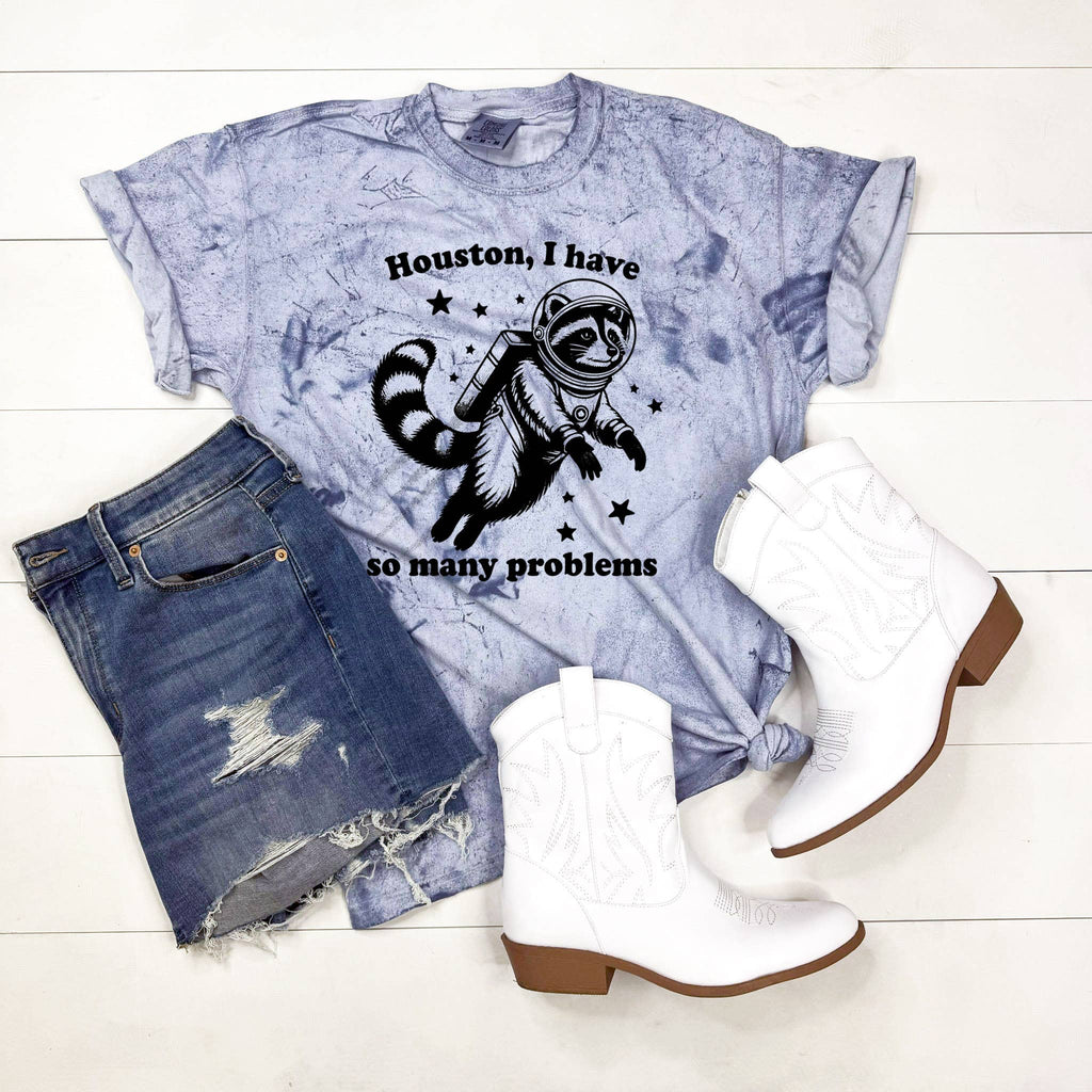 T-shirt with a raccoon astronaut graphic and text, paired with denim shorts and white boots on a white wooden surface.