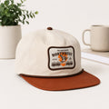 An off-white baseball cap with a brown flat brim and a front patch reading 'The Great Northwest' with an orange fish graphic, styled on a white surface with a potted plant and a mug in the background.