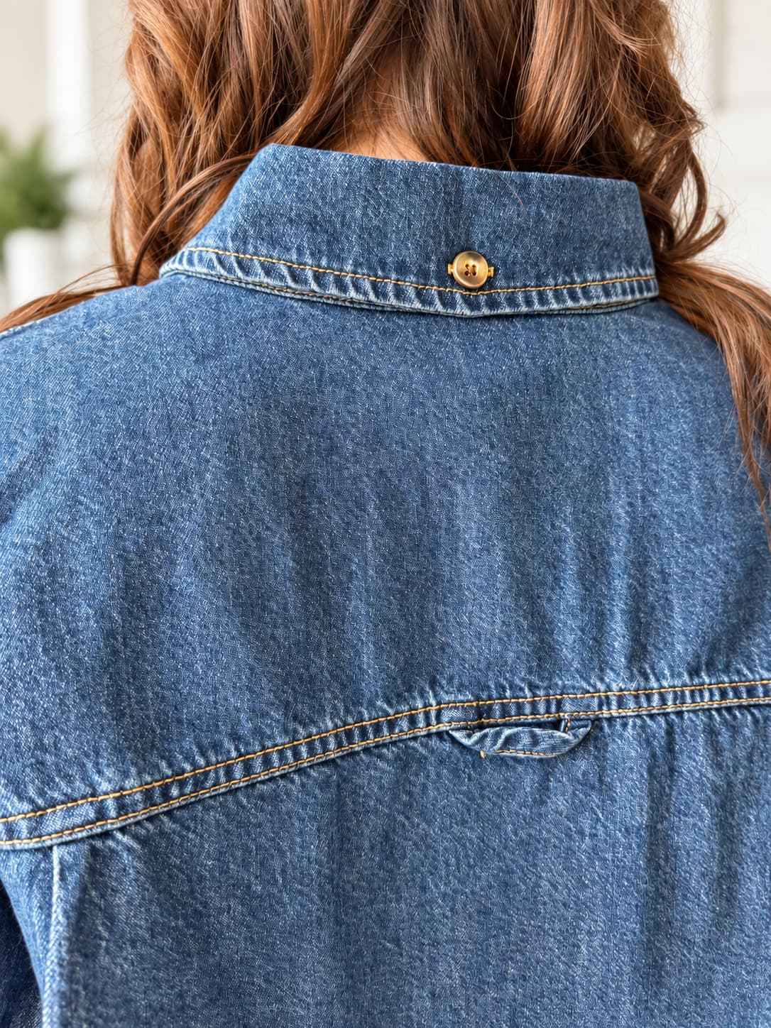 Close-up back view of a person wearing a blue denim shirt jacket, showing the collar, button, and back fabric details.