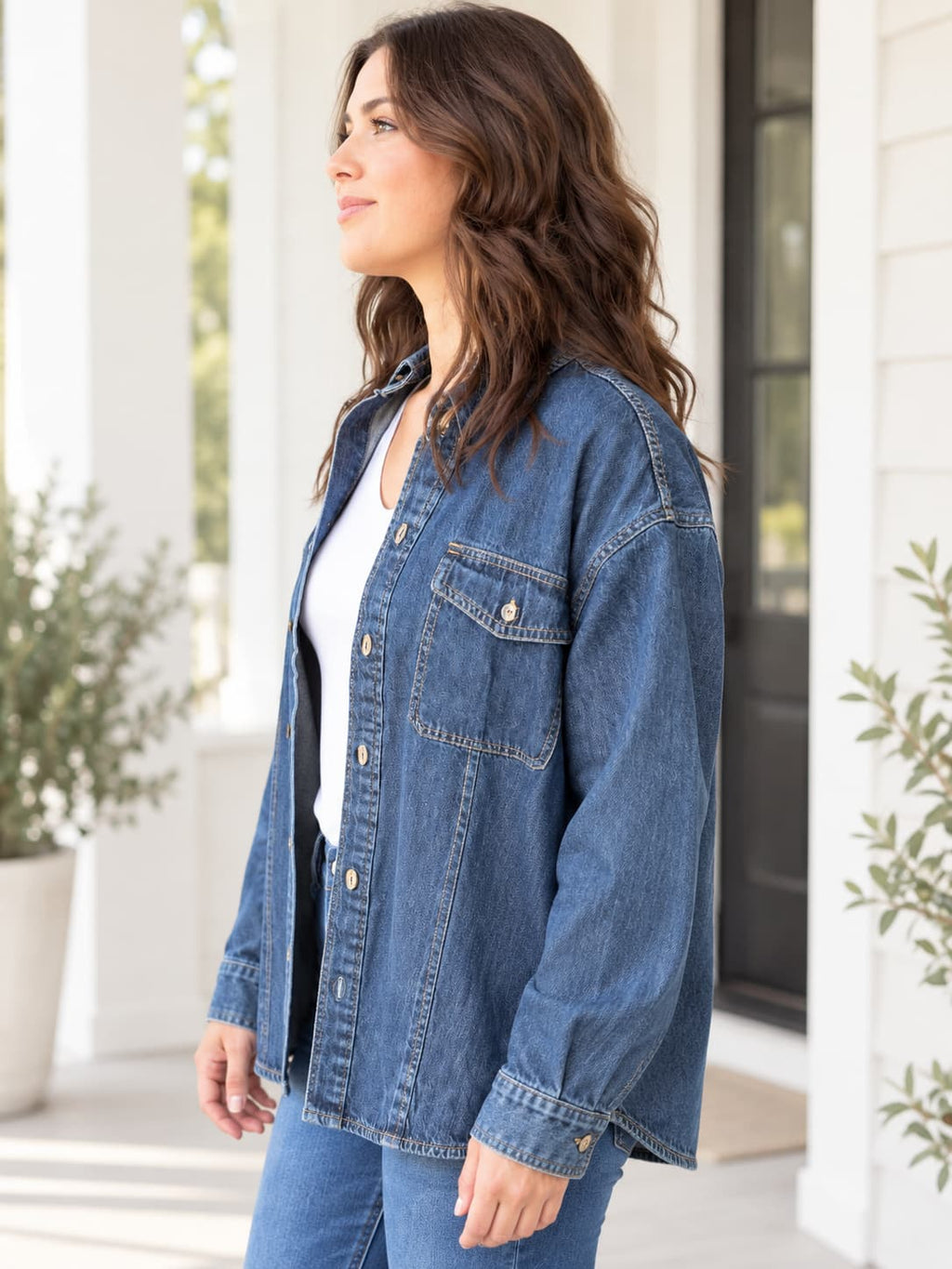 A woman wearing a blue denim button-up shirt jacket over a white top and blue jeans, standing on a porch with white columns and plants in the background.