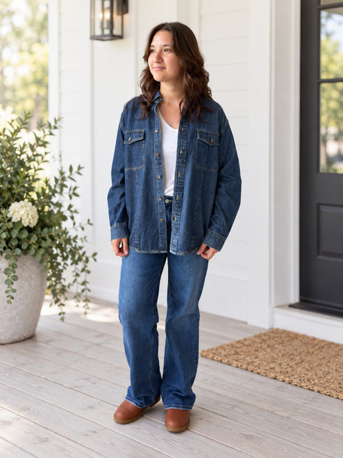 A woman stands on a porch wearing a blue denim button-up shirt and matching blue jeans over a white top, with brown shoes. The porch has white siding, a black door, a doormat, and a potted plant.