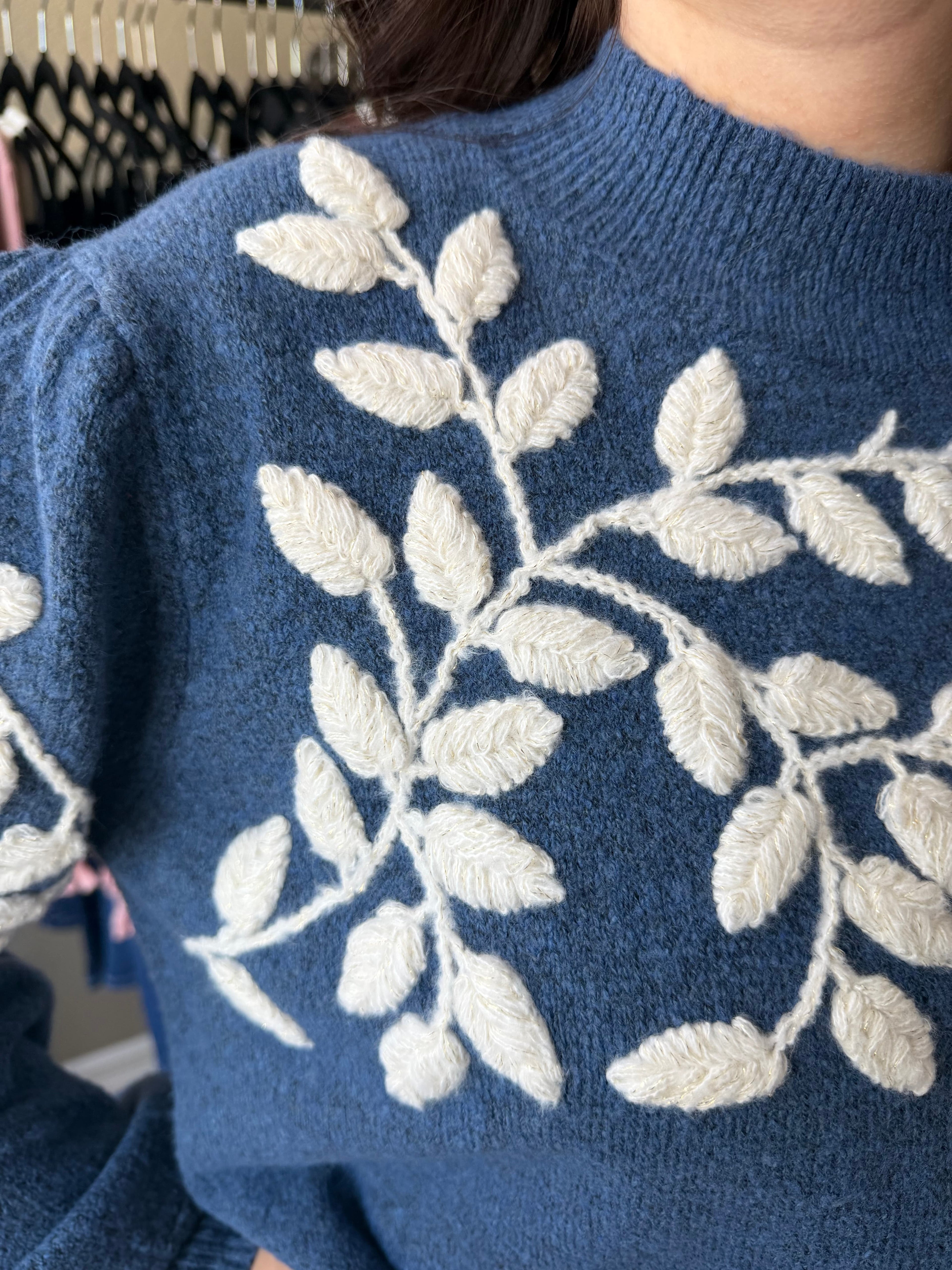Close-up of a blue knit sweater with a large white leafy branch  yarn appliqué  pattern across the chest and shoulders, worn by a person in an indoor setting.