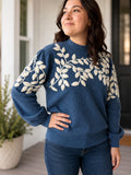 A woman wearing a navy blue knit sweater with cream-colored leafy branch yarn appliqué  patterns across the chest and shoulders, paired with dark blue jeans, standing in a brightly lit indoor setting.