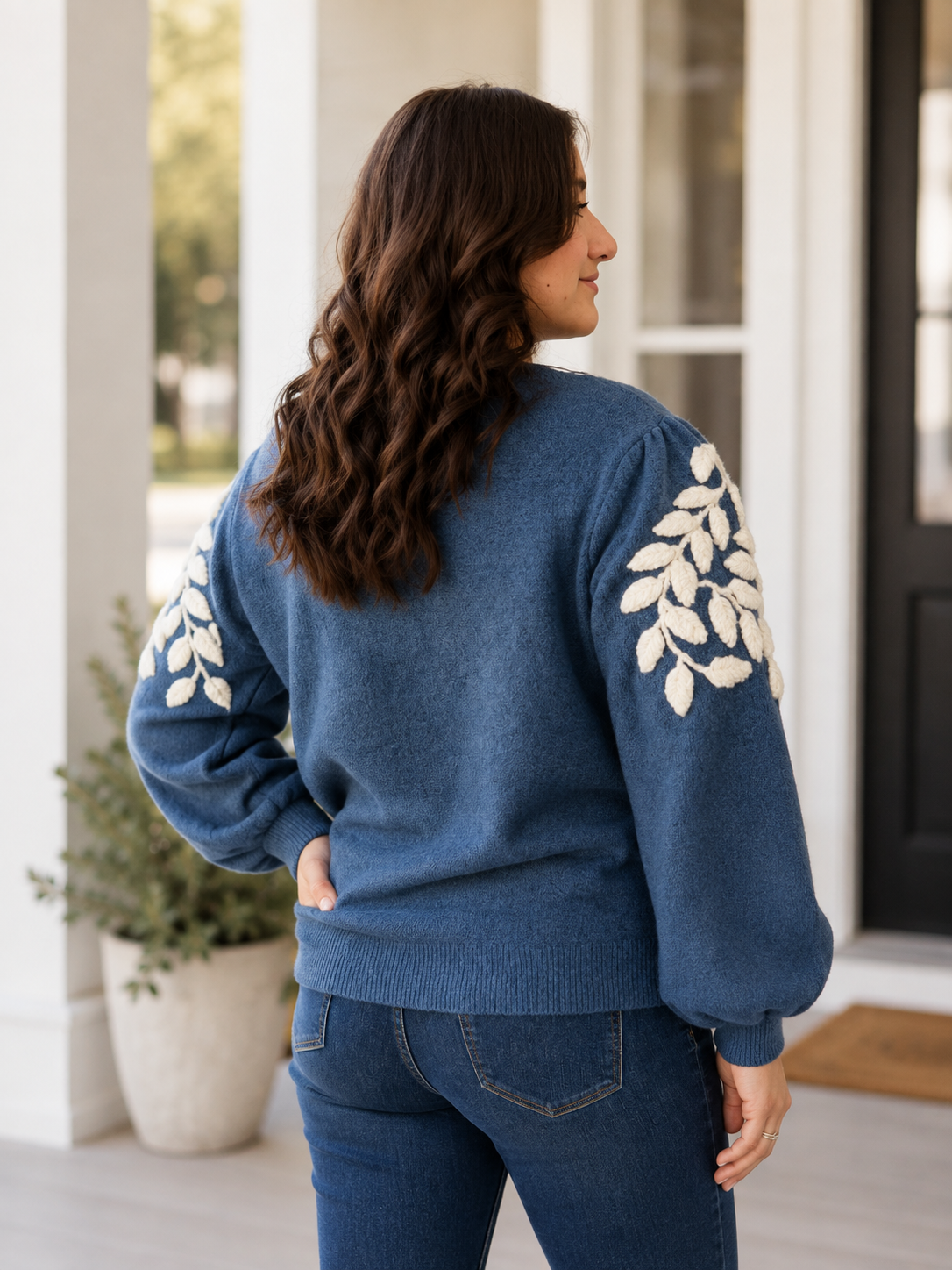 A woman wearing a blue knit sweater with cream leaf  yarn appliqué on the sleeves, shown from the back in an outdoor porch setting.
