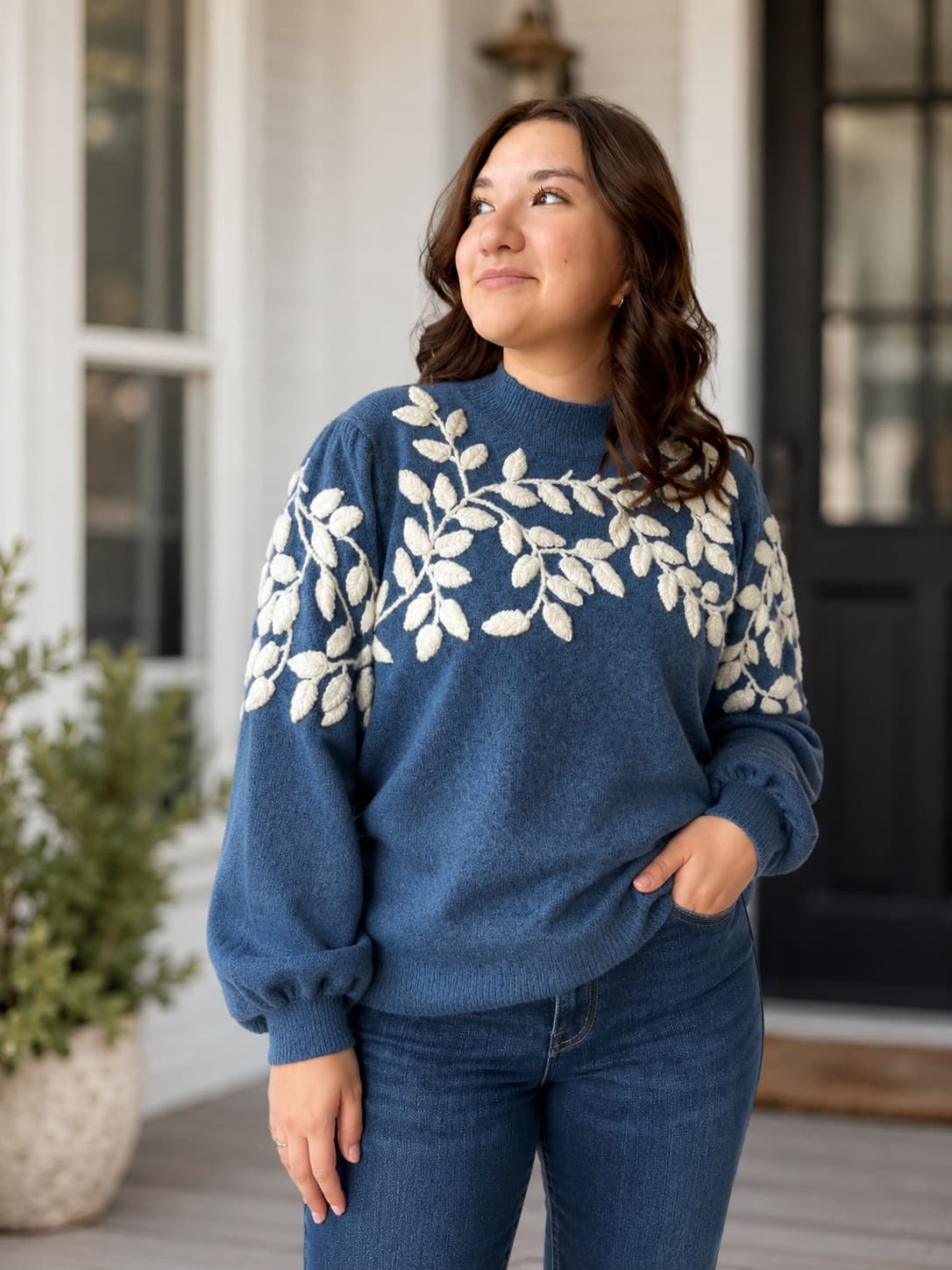 A woman wearing a blue crewneck sweater with a cream leafy vine yarn appliqué pattern across the chest and shoulders, paired with blue jeans, standing on a front porch.