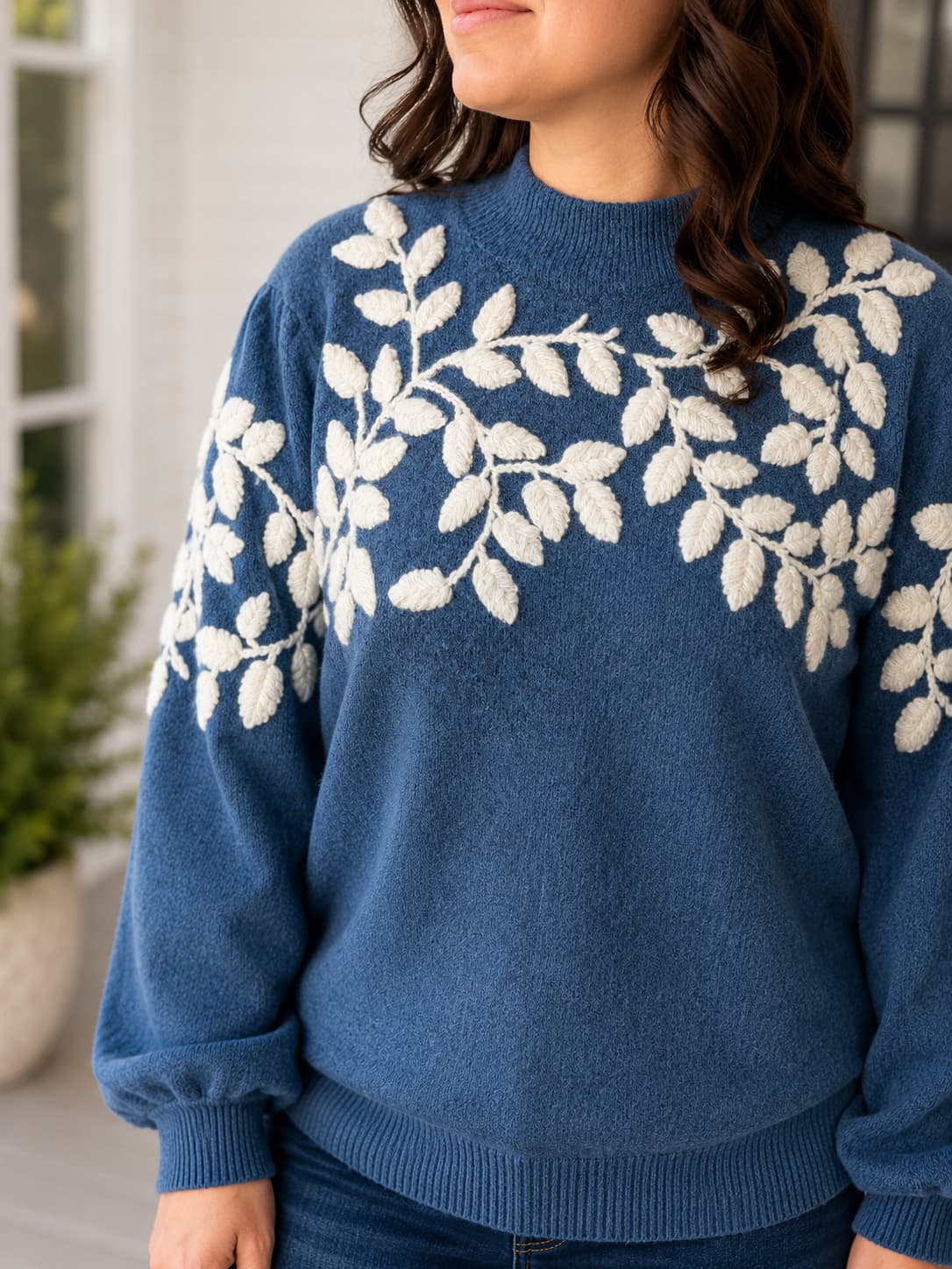 A woman wearing a blue crewneck sweater with a white leafy yarn appliqué  vine pattern across the chest and shoulders, paired with dark blue jeans.