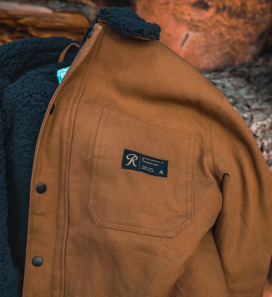 A close-up of a brown overshirt jacket with a dark navy fleece lining, featuring a chest pocket with a black brand label that reads 'R' and 'The Rough Draft Co.'.