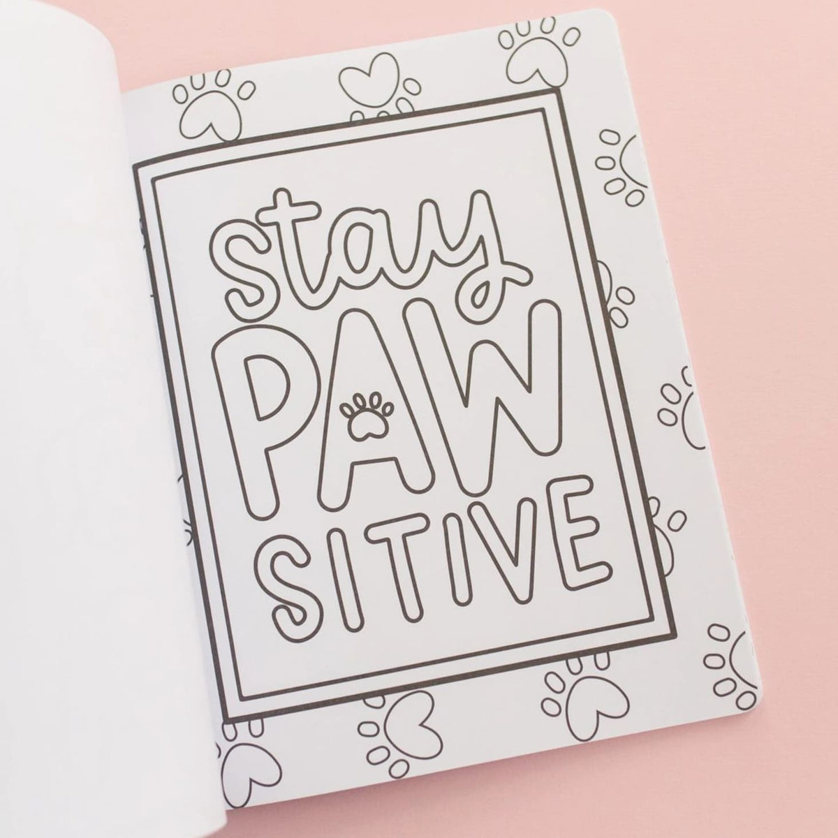 A white coloring book or notebook page with black outline drawings of paw prints and the phrase 'stay PAW SITIVE' in a bordered box, photographed on a soft pink background.