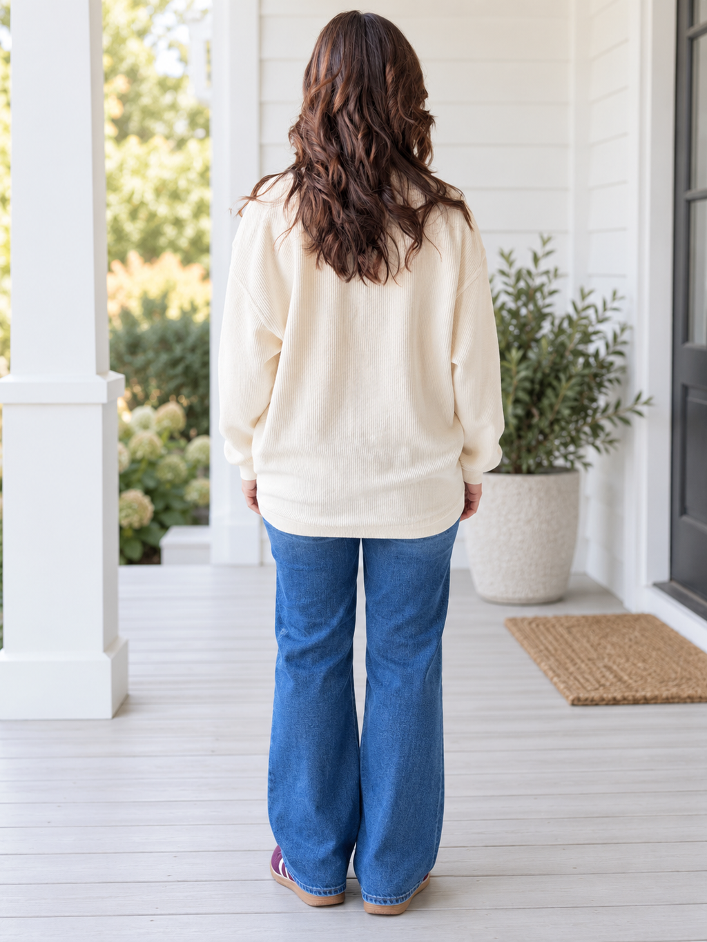 A woman viewed from behind wearing a cream-colored long-sleeve knit top and blue wide-leg jeans, standing on a white porch with a potted plant in the background.