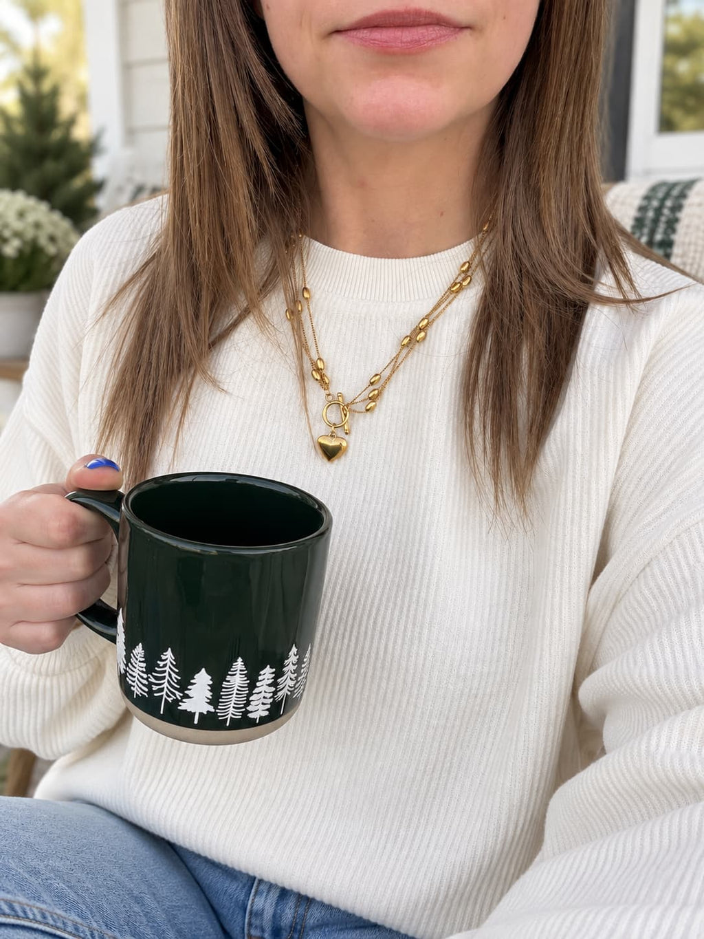 A woman wearing a white ribbed knit sweater and gold chain necklace holds a dark green ceramic mug with a white pine tree pattern, set against a softly blurred indoor background.