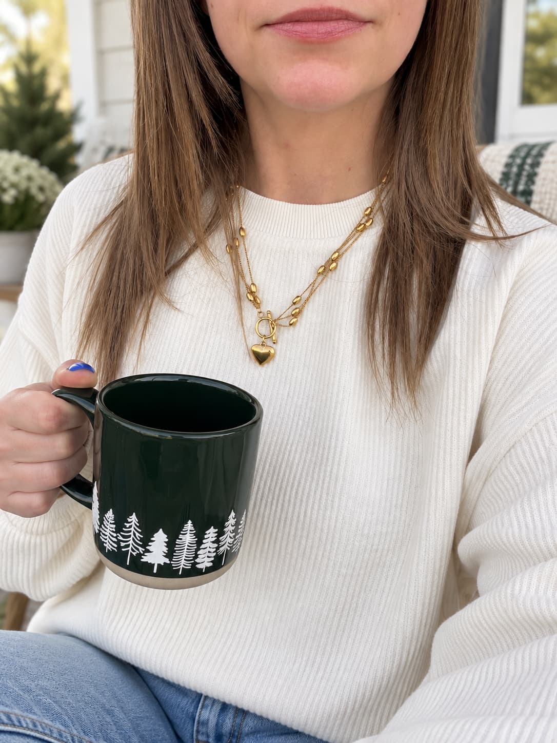 A woman wearing a white ribbed knit sweater and gold chain necklace holds a dark green ceramic mug with a white pine tree pattern, set against a softly blurred indoor background.
