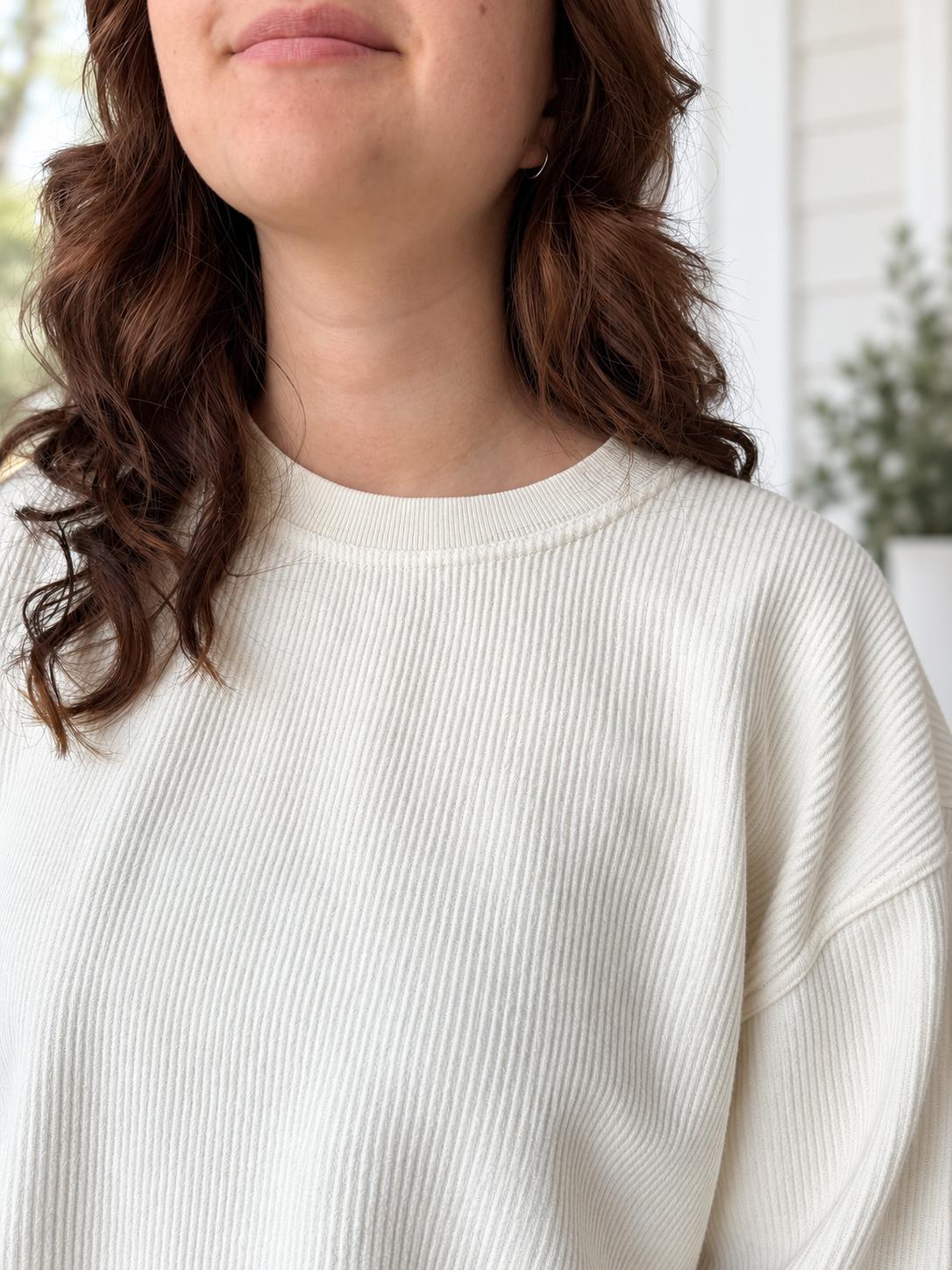 Close-up of a woman wearing a ribbed cream-colored long-sleeve top, showing her hair, neck, and upper chest against a softly blurred light background.