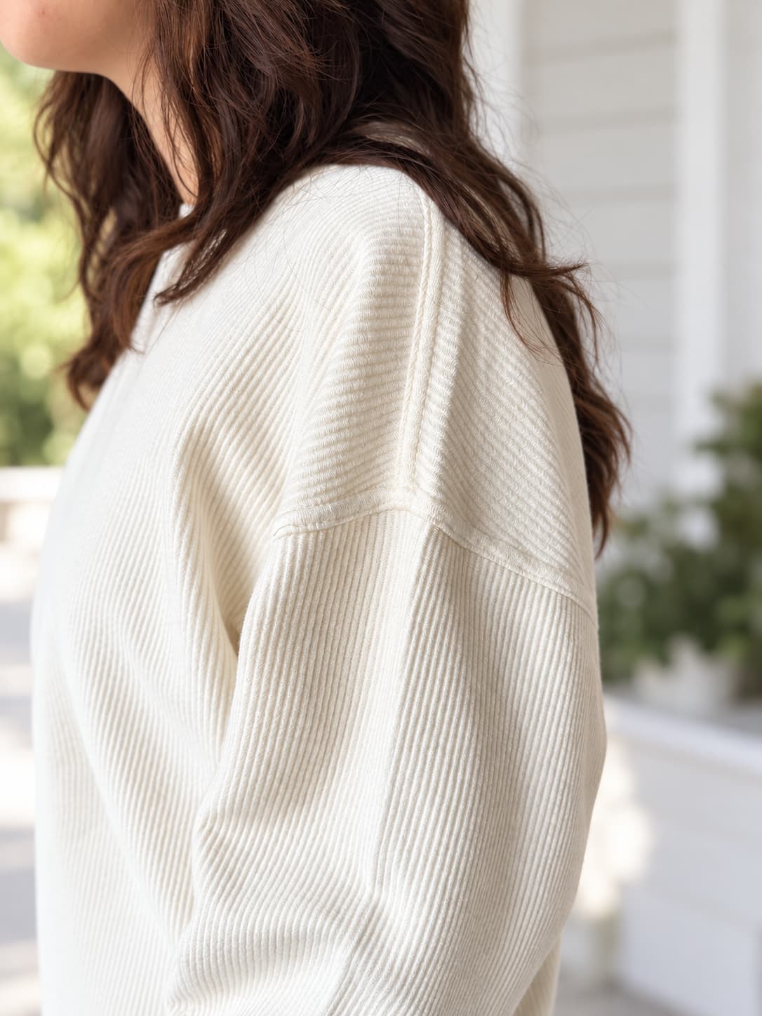 A close-up side view of a woman wearing a cream-colored ribbed knit sweater, with her brown hair visible and a softly blurred porch background.