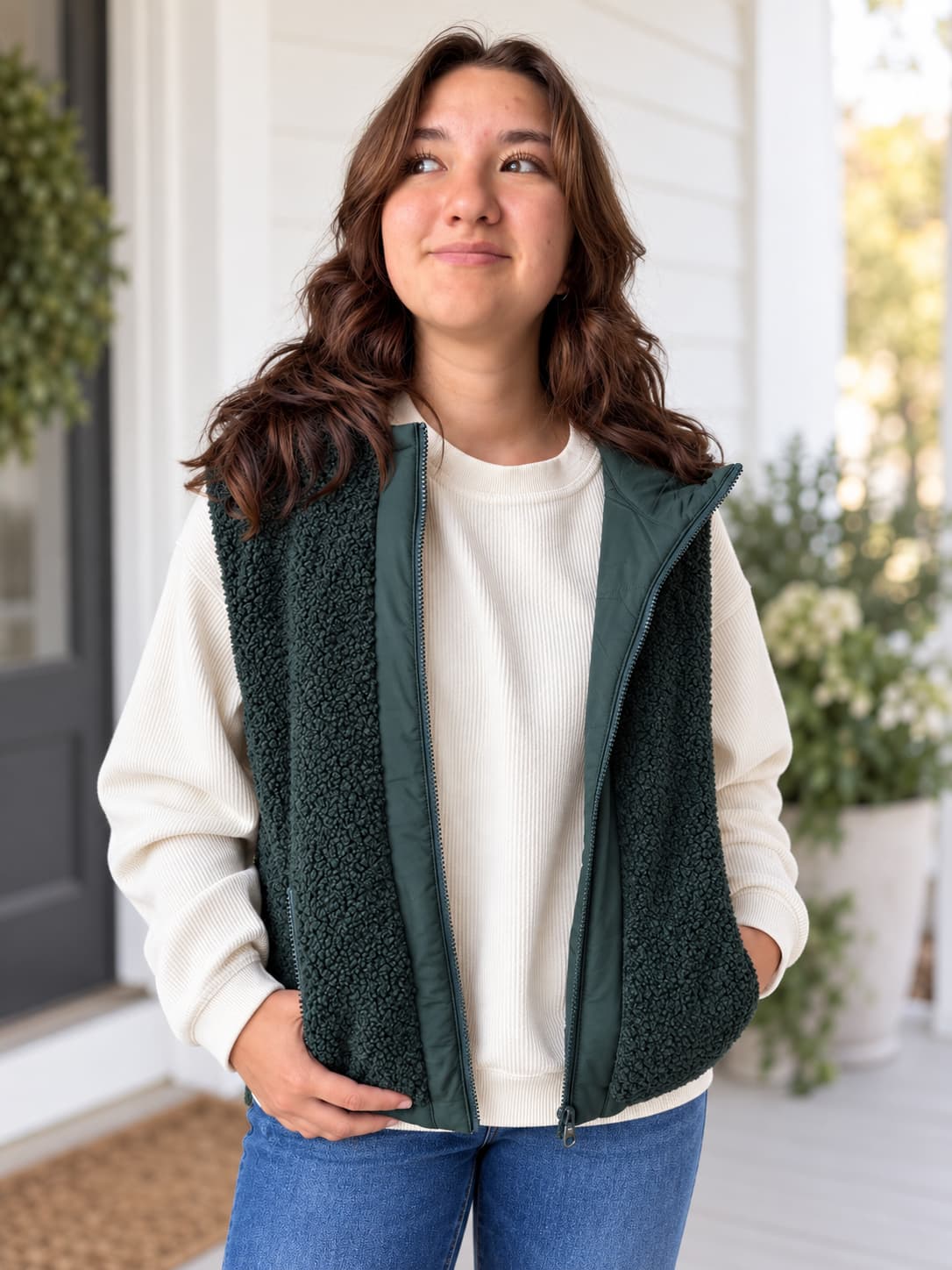 A woman wearing a dark green fleece vest over a cream ribbed sweatshirt and blue jeans, standing on a front porch.