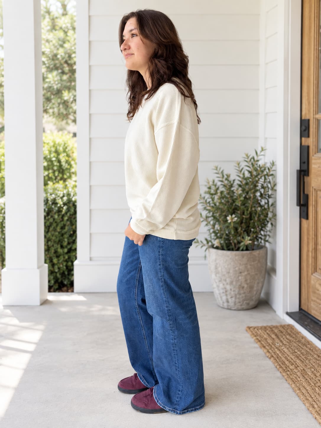 A woman stands in profile on a porch wearing a cream long-sleeve sweatshirt, wide-leg blue jeans, and burgundy slip-on shoes. The background shows white house siding, a potted plant, and a wooden door.