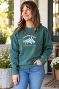 A woman wearing a dark teal green crewneck sweatshirt with a graphic that reads 'WASHINGTON THE EVERGREEN STATE' above a mountain and deer design, paired with blue jeans, standing on a porch.