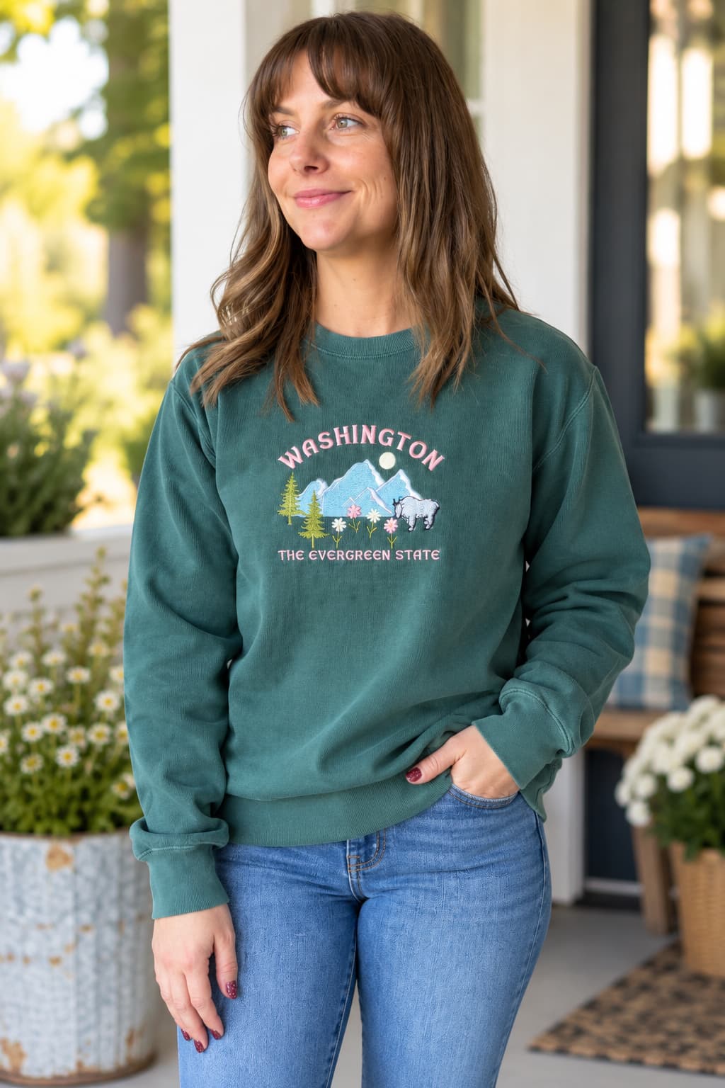 A woman wearing a dark teal green crewneck sweatshirt with a graphic that reads 'WASHINGTON THE EVERGREEN STATE' above a mountain and deer design, paired with blue jeans, standing on a porch.