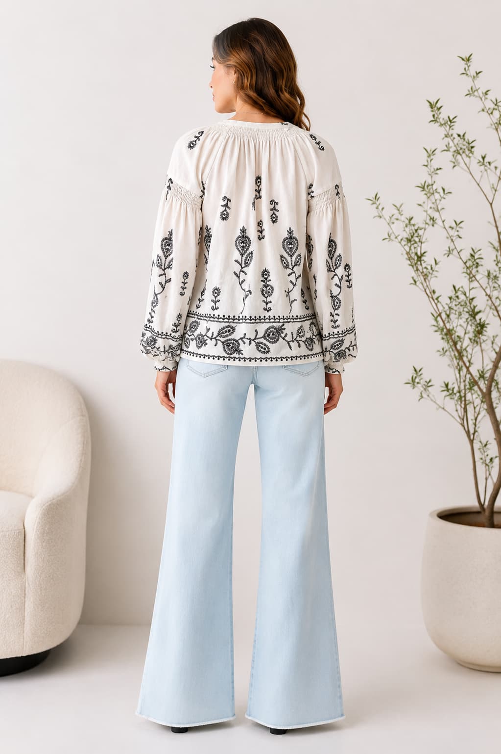 Back detail of woman wearing a white blouse with black embroidery and light blue jeans in a minimalistic room.