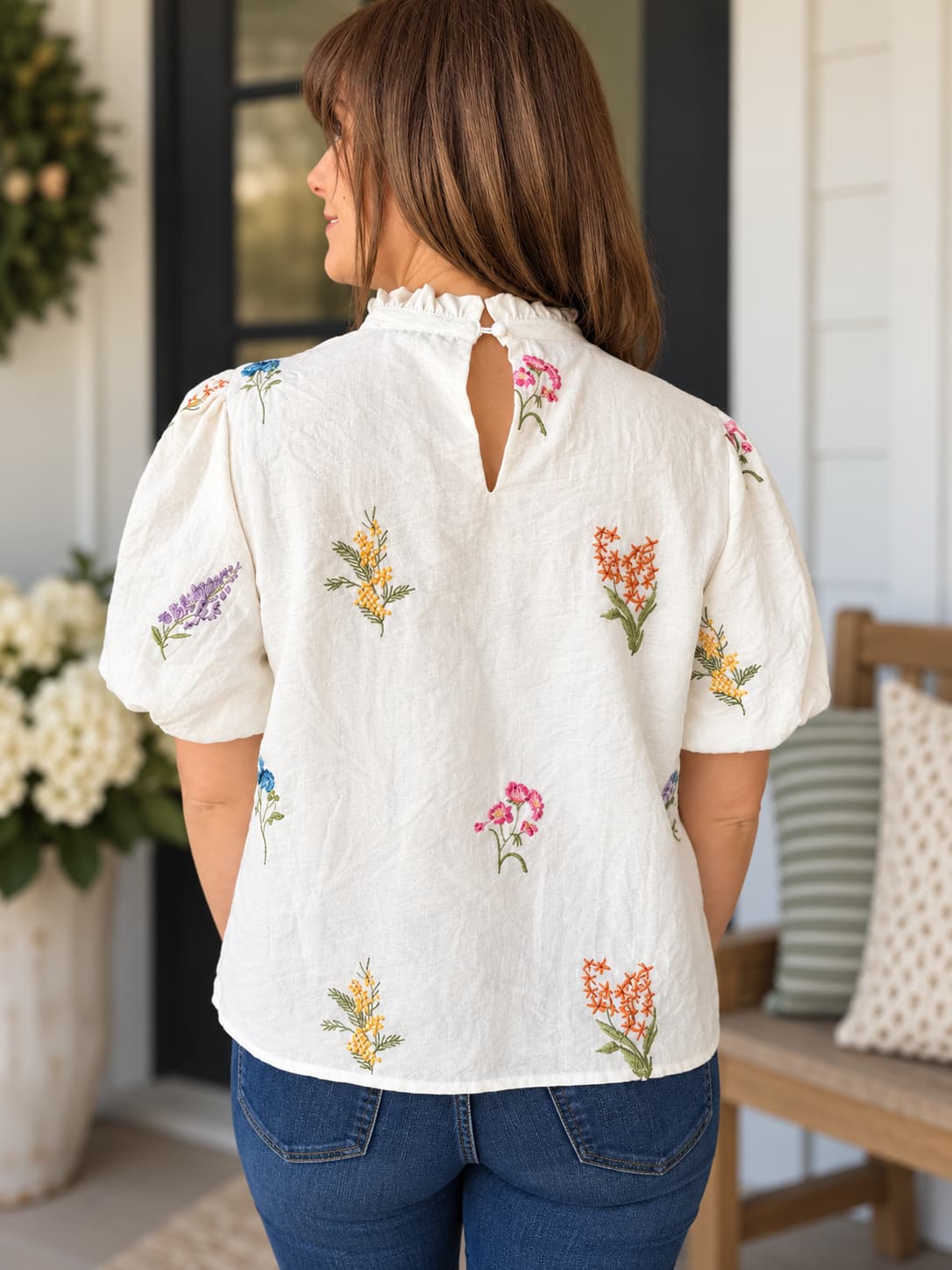A woman viewed from the back wearing a white short-sleeve blouse with colorful floral embroidery and a small keyhole cutout at the neckline, paired with blue jeans in a porch setting.