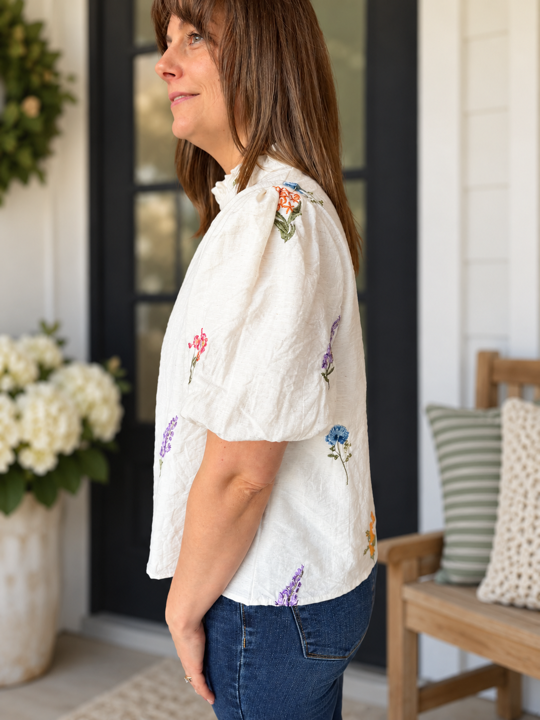 A woman wearing a white short-sleeve blouse with colorful floral embroidery and blue jeans, standing on a porch.