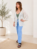 Woman wearing a white lace kimono-style top and blue jeans standing in a bright room with a plant in the background.