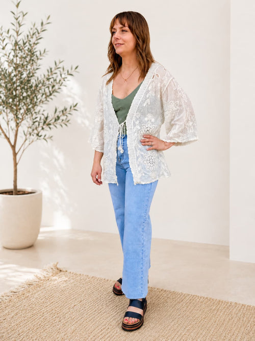 Woman wearing a white lace kimono-style top and blue jeans standing in a bright room with a plant in the background.