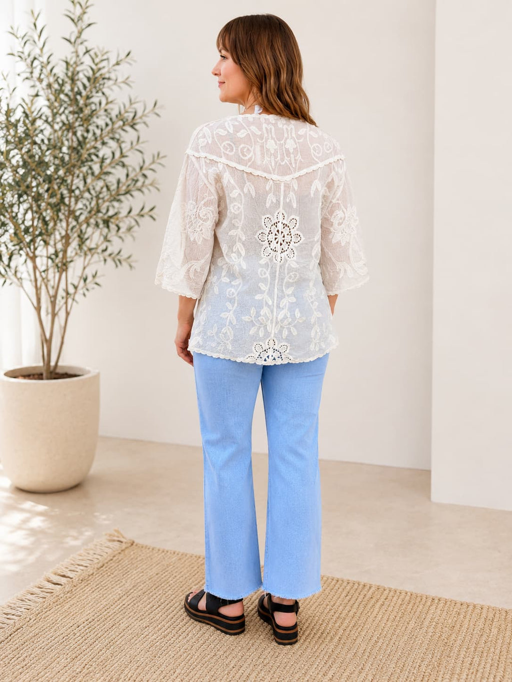 Woman wearing a white embroidered kimono top and blue pants standing in a room with a plant and rug.