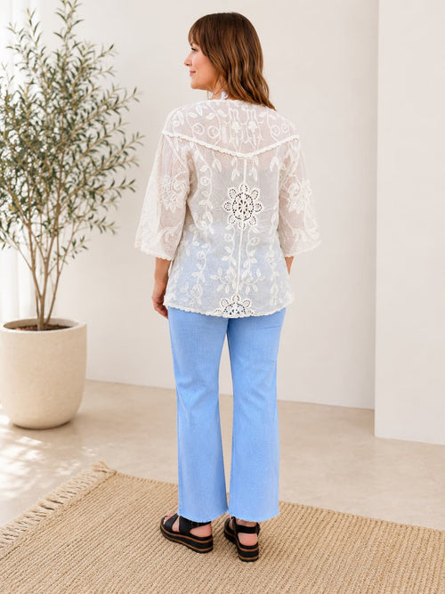 Woman wearing a white embroidered kimono top and blue pants standing in a room with a plant and rug.