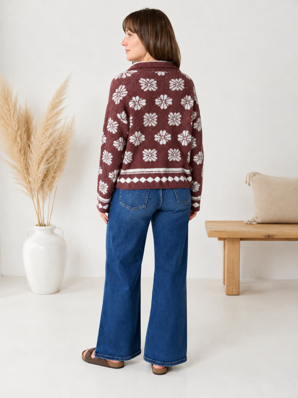A woman shown from the back wearing a brown knit cardigan with a white Nordic snowflake pattern, paired with blue wide-leg jeans and brown sandals, standing in a bright room with a pampas grass vase and a wooden bench.