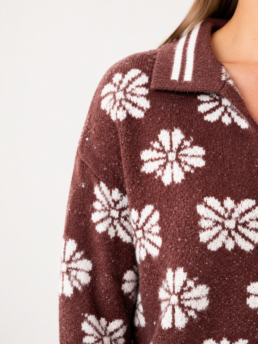 Close-up of a person wearing a brown knit collared sweater with a white floral pattern and white stripes on the collar, against a plain light background.