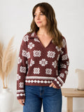 A woman wearing a burgundy knit sweater with a white Nordic snowflake pattern and blue jeans, standing in a brightly lit room with a wooden bench and pampas grass in the background.