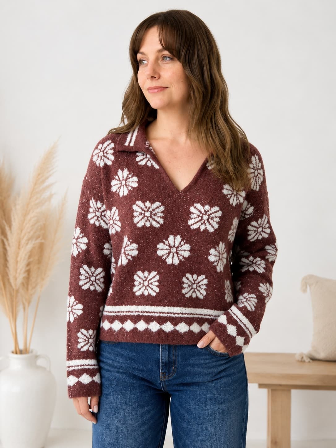 A woman wearing a burgundy knit sweater with a white Nordic snowflake pattern and blue jeans, standing in a brightly lit room with a wooden bench and pampas grass in the background.